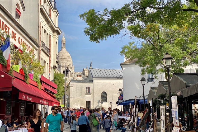 crowds in a street with artist stalls and cafes and one of the domes of sacre-couer in the background
